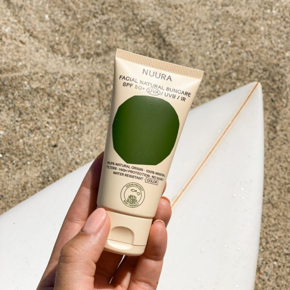 Mano sosteniendo la crema solar facial fluida con color de Nuura en la playa junto a una tabla de surf