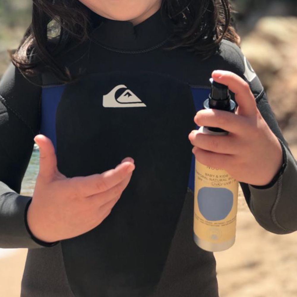 Niño con neopreno en la playa sosteniendo el protector solar mineral Nuura Kids SPF50 para mostrar su cómodo formato con dosificador.