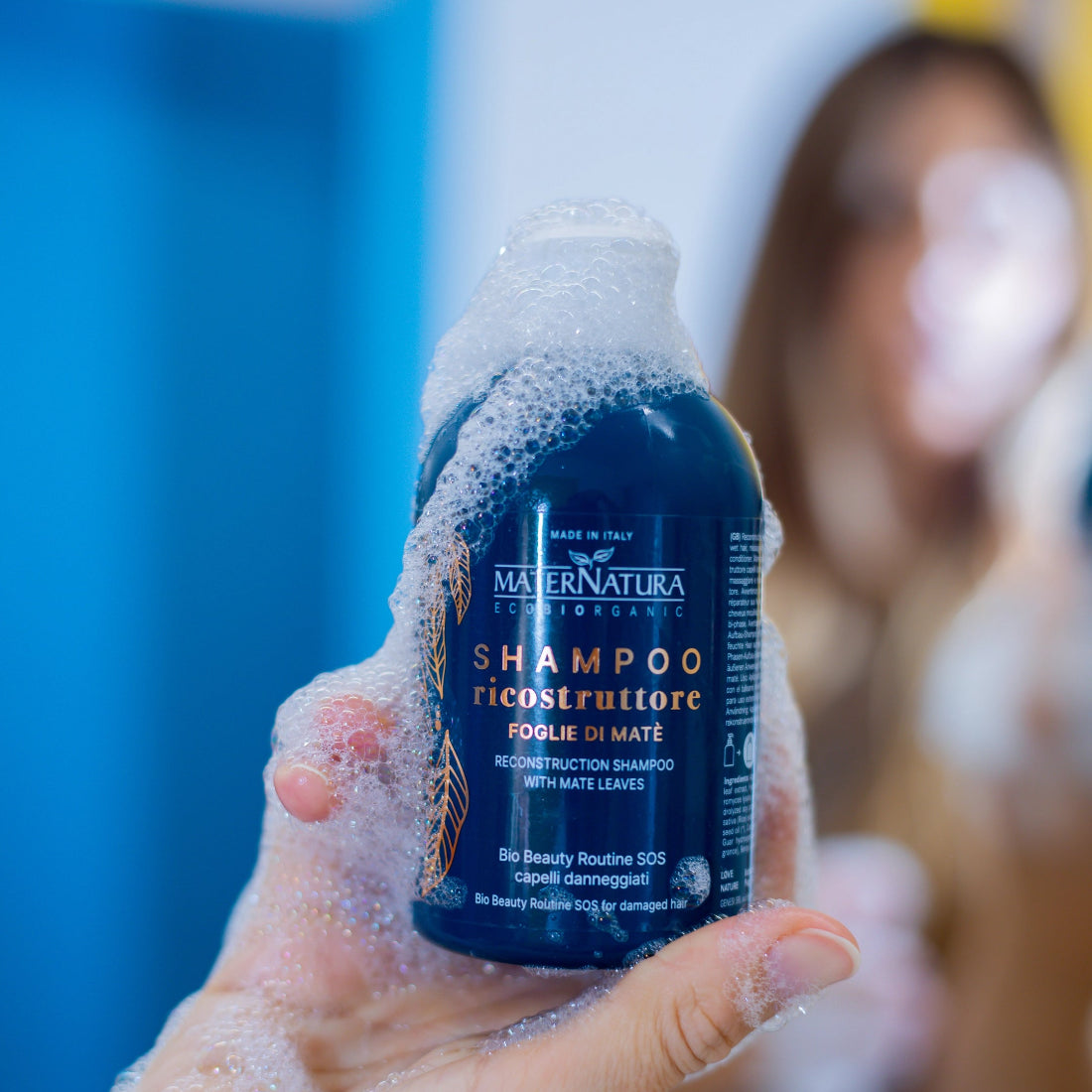 Una mano sostiene el Champú Reconstructor de MaterNatura cubierto de espuma cremosa en el baño, con una mujer desenfocada al fondo.