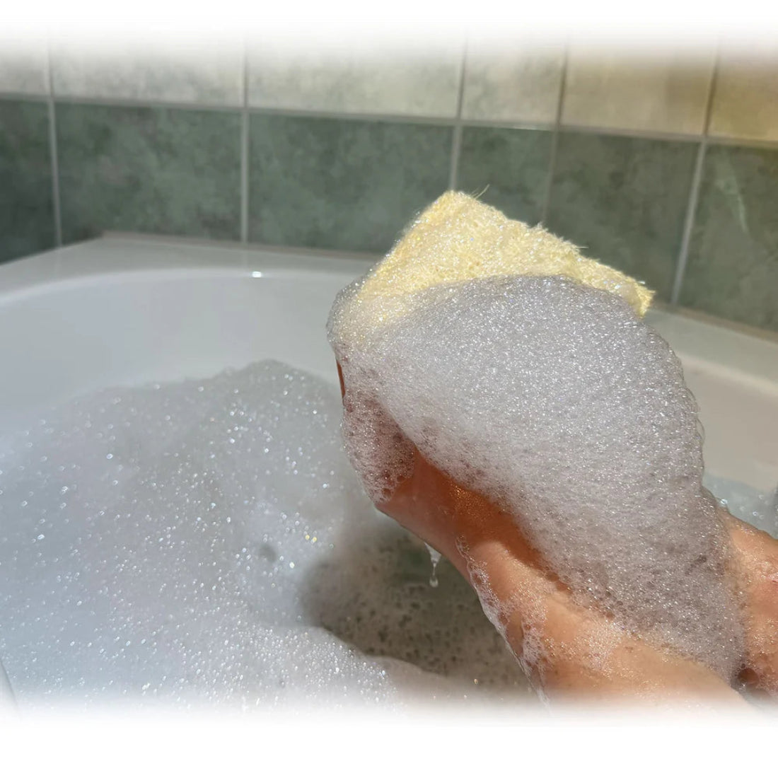 Primer plano de una mano sosteniendo la esponja de lufa natural de Iberluffa cubierta de abundante espuma blanca dentro de una bañera