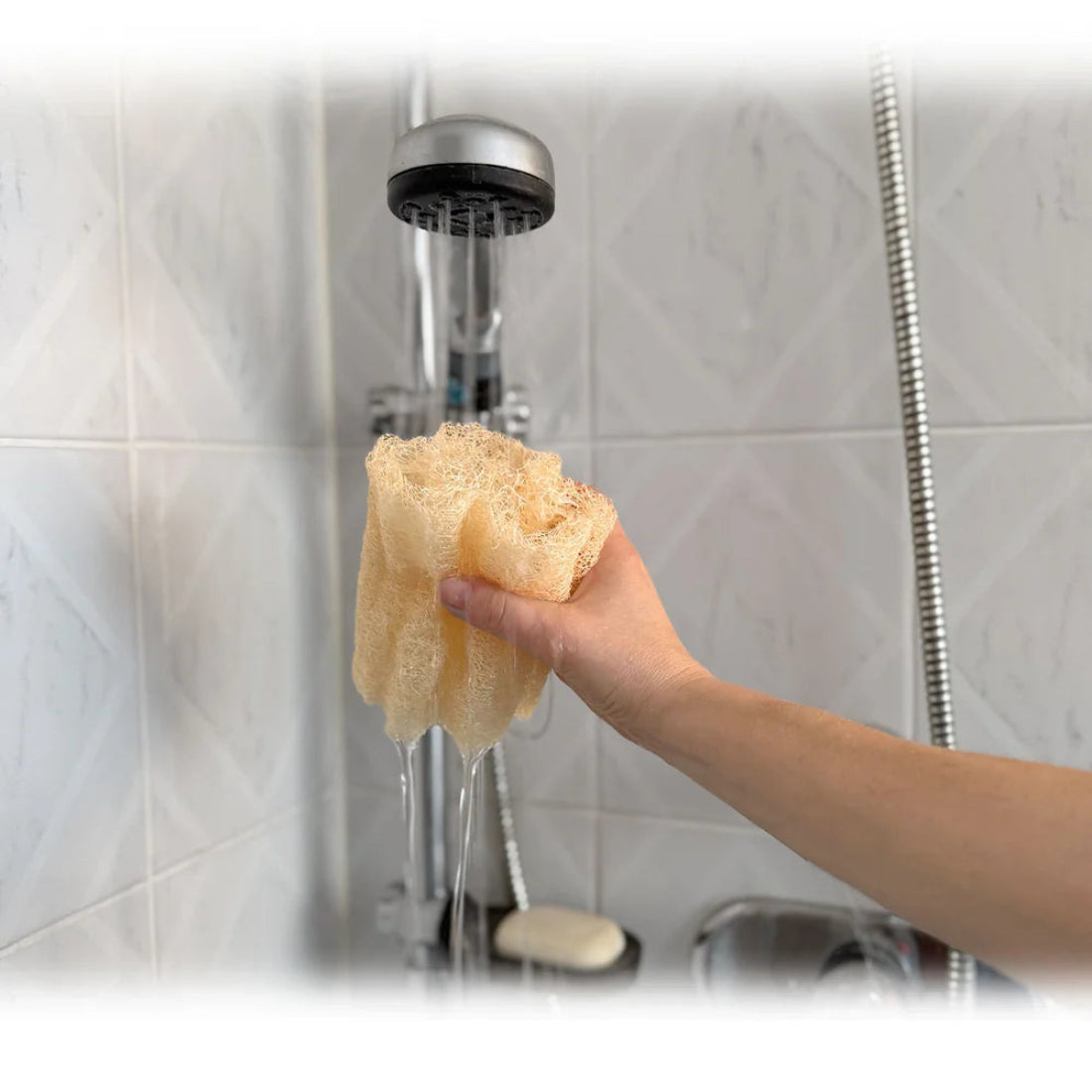 Mano sosteniendo una esponja vegetal de luffa mojada bajo el agua de la ducha de una baño.