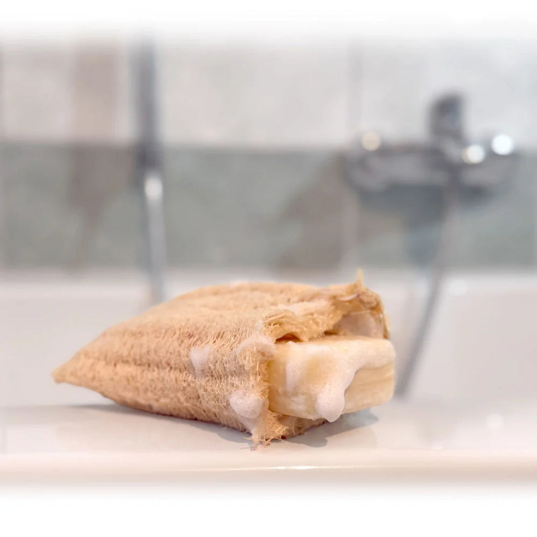 Funda de lufa natural con una pastilla de jabón sólido en su interior generando abundante espuma blanca en el borde de una bañera.