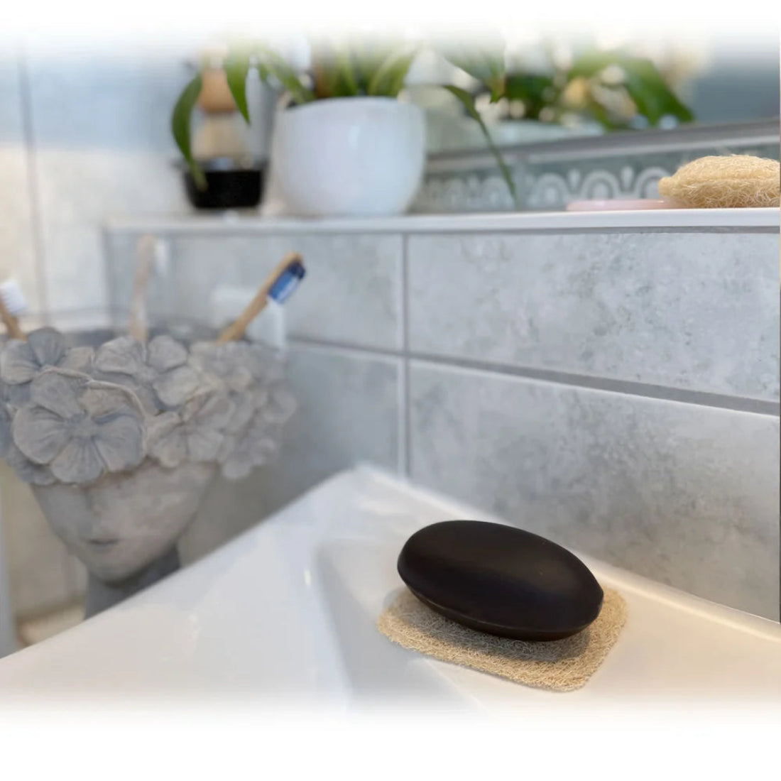 Jabón sólido negro apoyado sobre un posajabones de luffa natural en el lavabo del baño